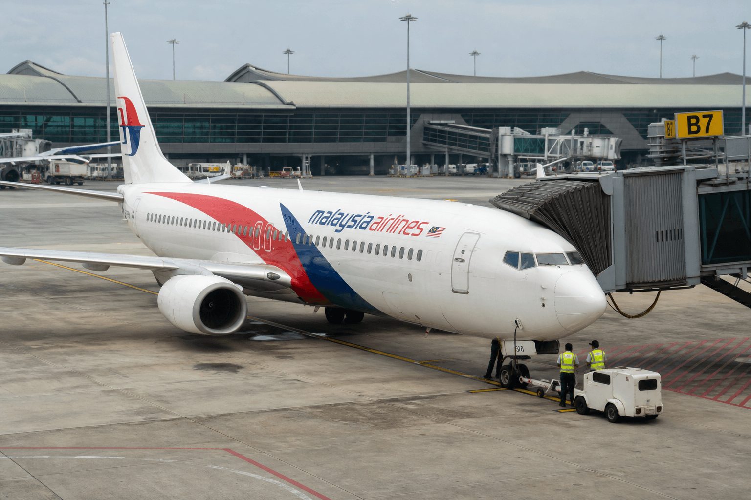 Malaysia Airlines aircraft parked at Kuala Lumpur International Airport with crew members on boarding bridge in daylight.
