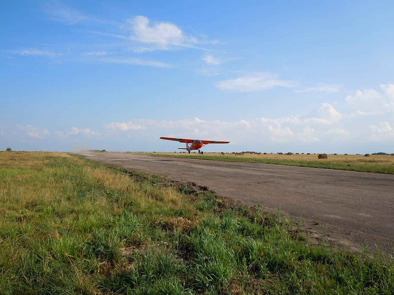 A Bell 407 helicopter hovering near a small single-engine Cessna 150 at a grass-lined runway in daylight